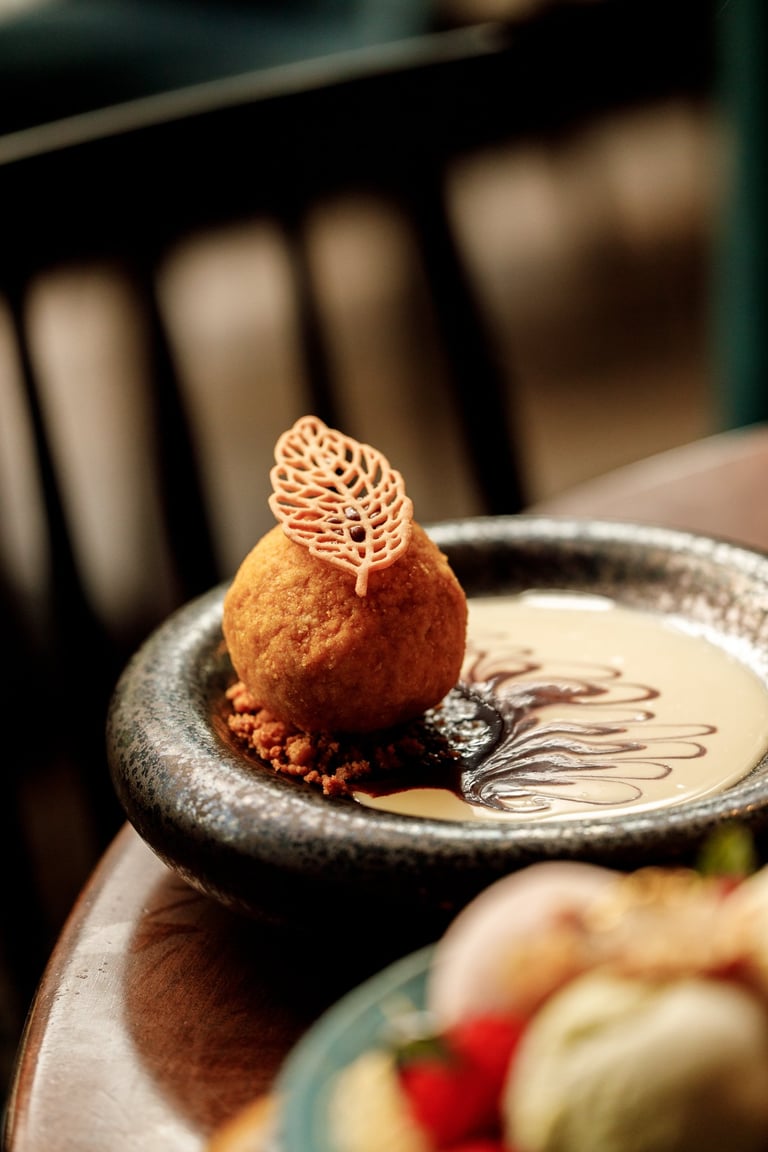 Artfully plated dessert featuring a golden fried sphere with delicate lattice tuile on top, served on a dark plate with chocolate sauce