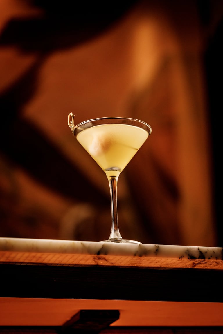 A pale yellow cocktail in a martini glass with an olive garnish, set on a wooden bar against a blurred warm brown background