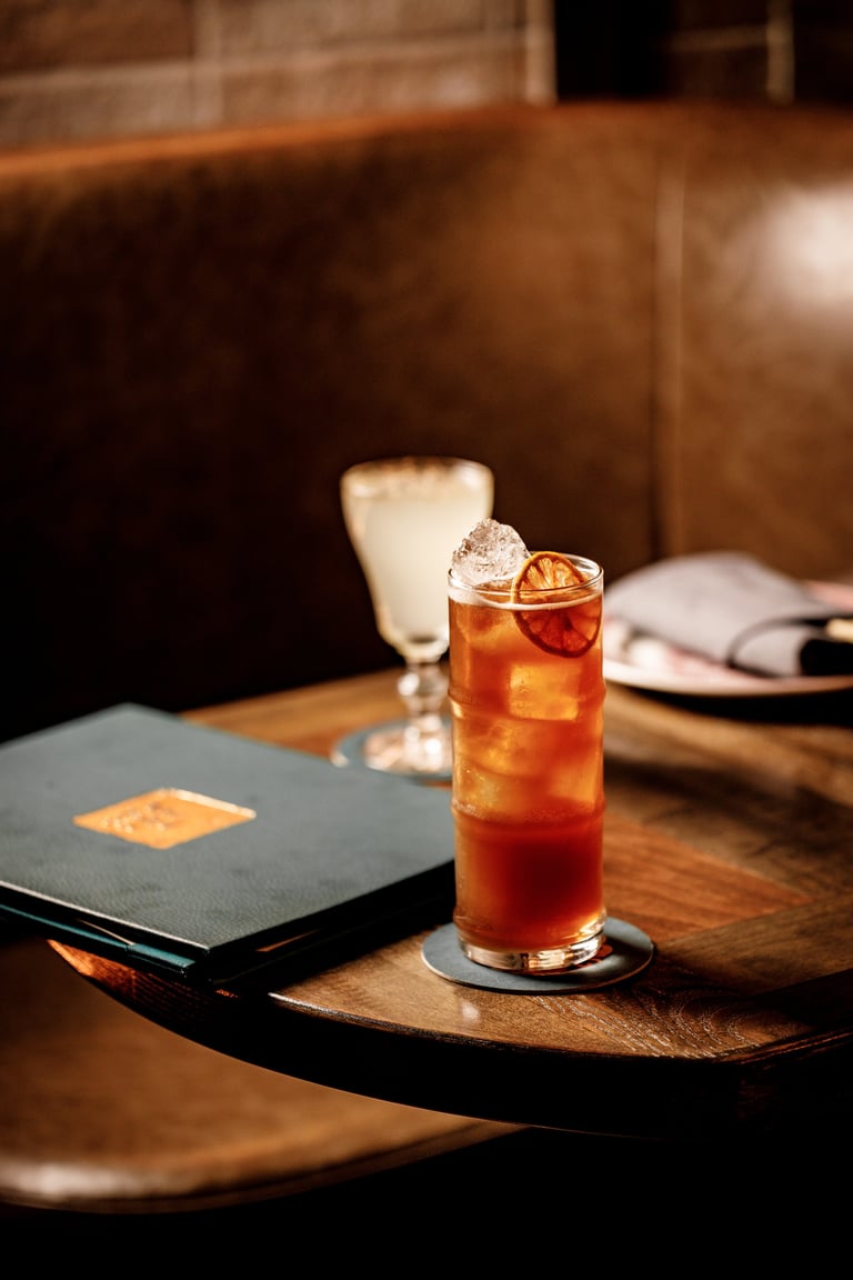 A glass of cola with ice and lemon slice on a wooden table beside a black notebook in an upscale lounge setting